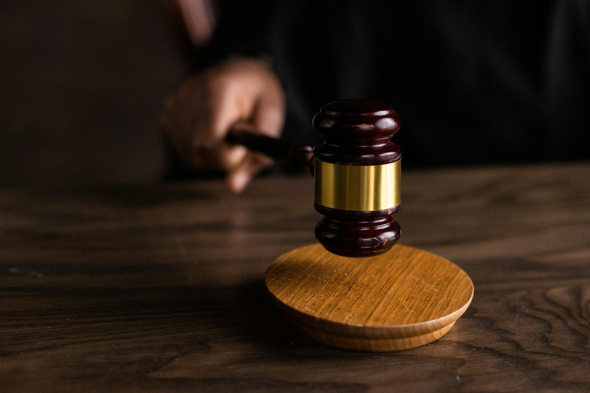 A hand holds a wooden gavel above a round sound block on a wooden table, suggesting a judicial or legal setting.