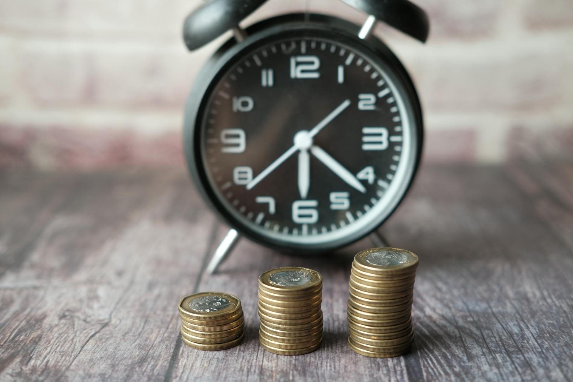 A black alarm clock and three stacks of coins are arranged on a wooden surface, with the coin stacks increasing in height from left to right.