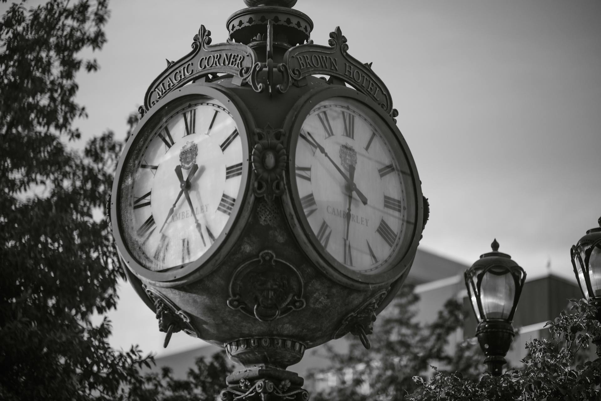 Black and white photo of a vintage double-faced street clock with Roman numerals, labeled “Magic Corner” and “Brown Hotel,” with trees and a streetlamp in the background.