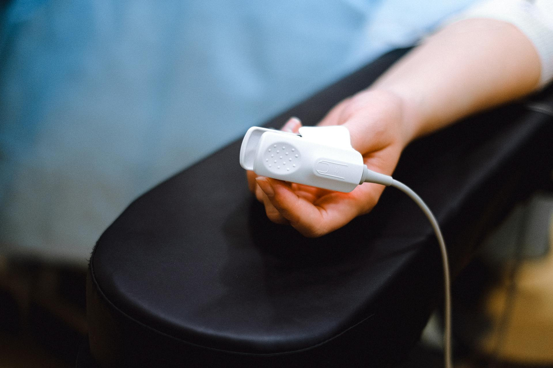 A person’s hand rests on a black armrest with a white pulse oximeter clip attached to their finger.