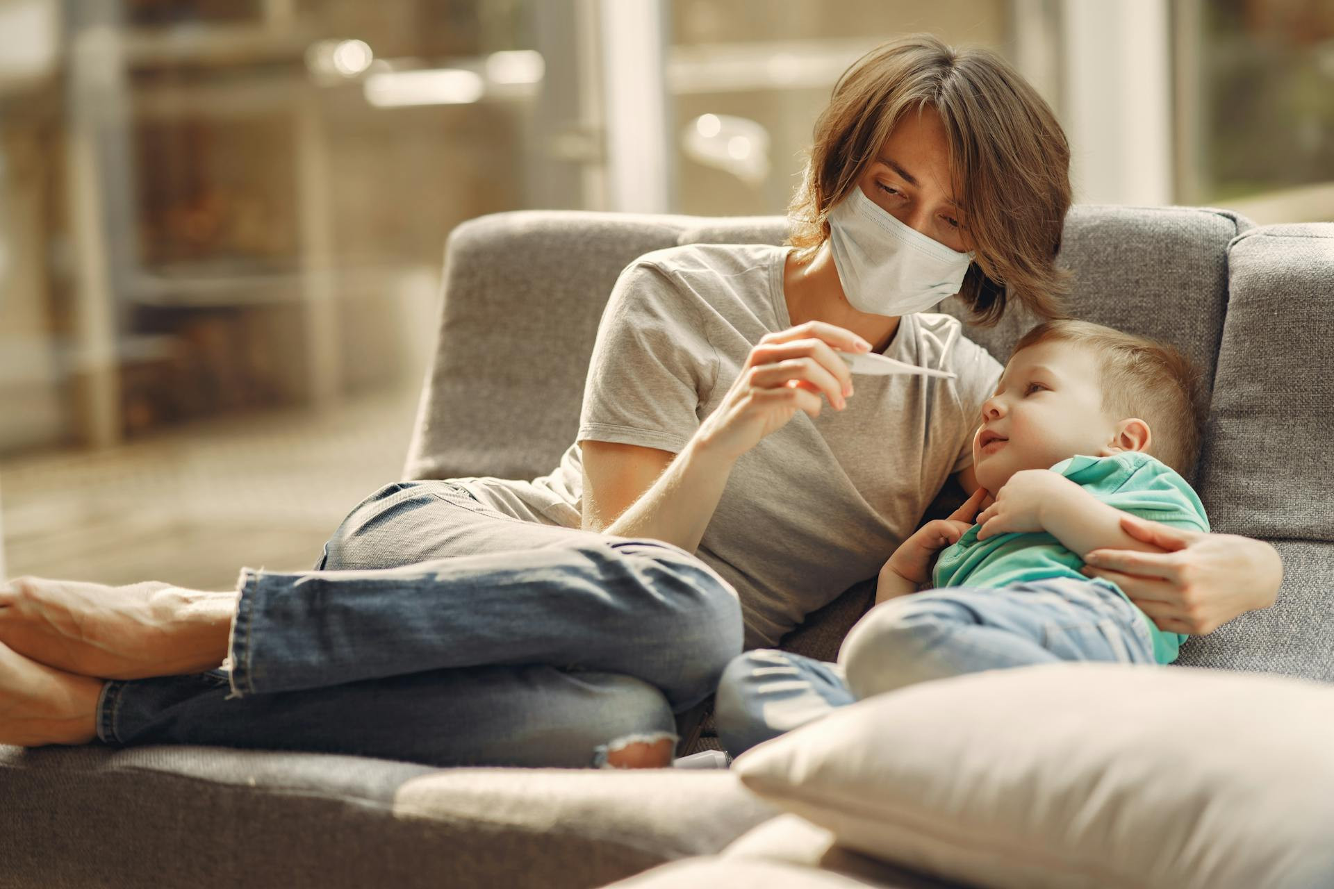 An adult wearing a face mask checks a thermometer while sitting on a couch with a young child lying beside them.