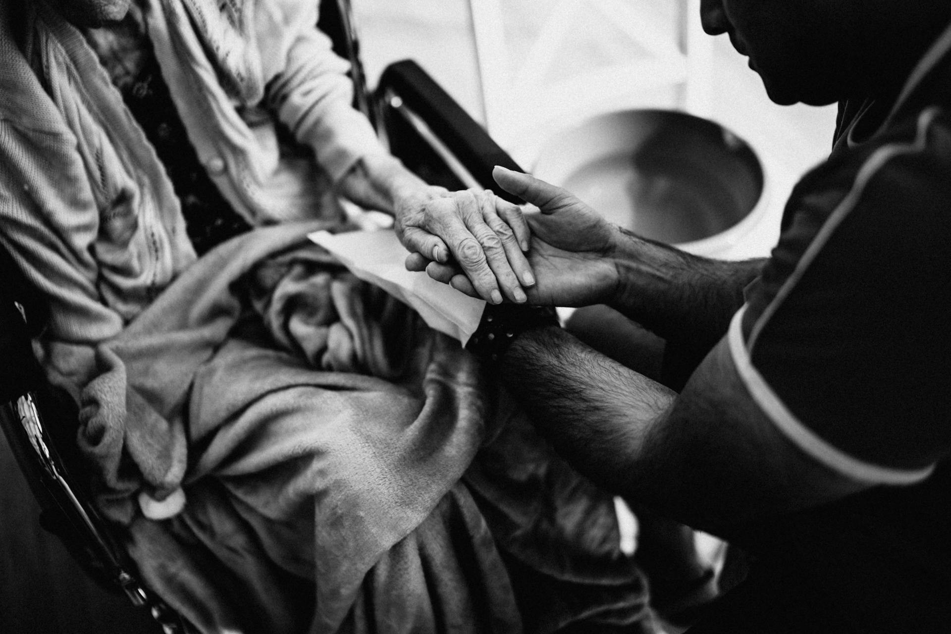 A person sitting in a wheelchair with a blanket is having their hand gently held and examined by another person.
