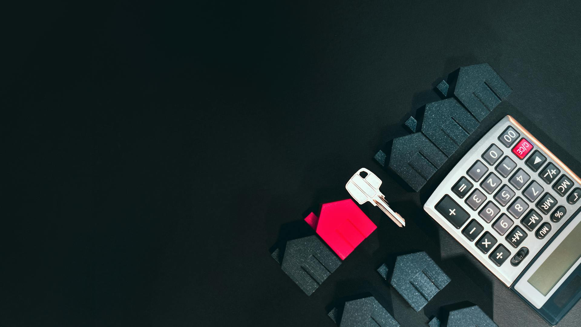 A calculator, a key, and several house-shaped figures, with one house in red, arranged on a dark surface.
