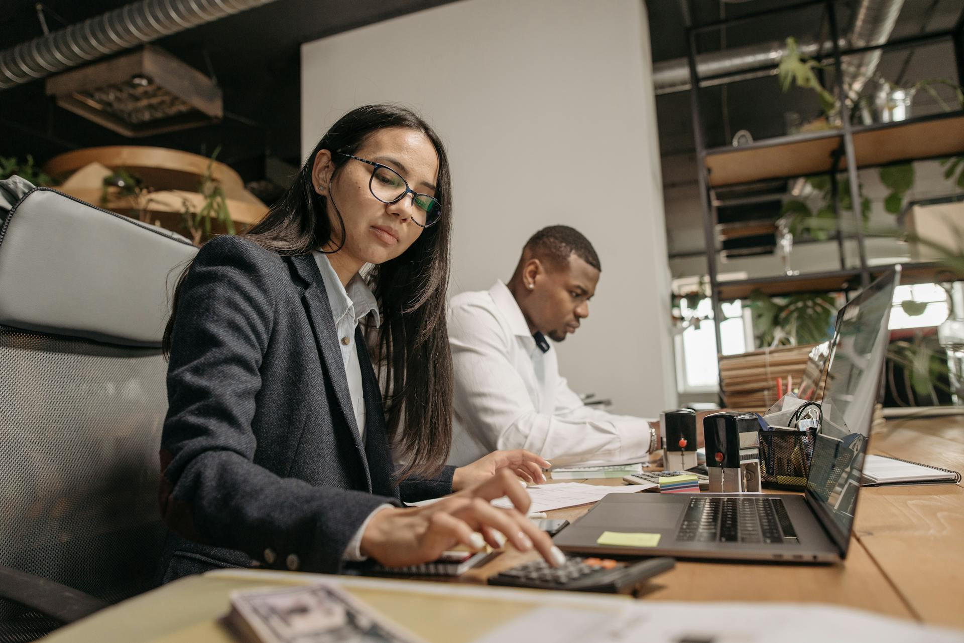 Two people sit at a desk working; the woman in front uses a calculator and a laptop, while the man beside her writes on paper. The office has plants and exposed pipes.
