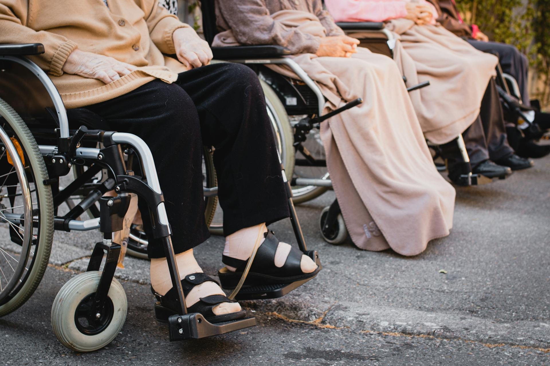 Four elderly people in wheelchairs sit side by side outdoors, with blankets covering their laps and legs.