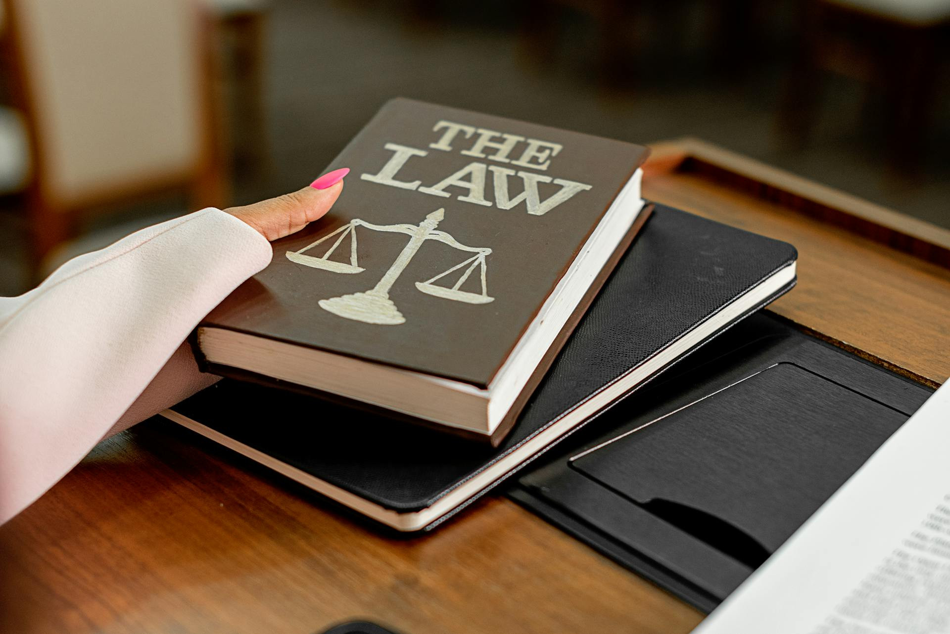 A person’s hand places a book titled "The Law" with scales of justice symbol on a desk, next to a closed black notebook.