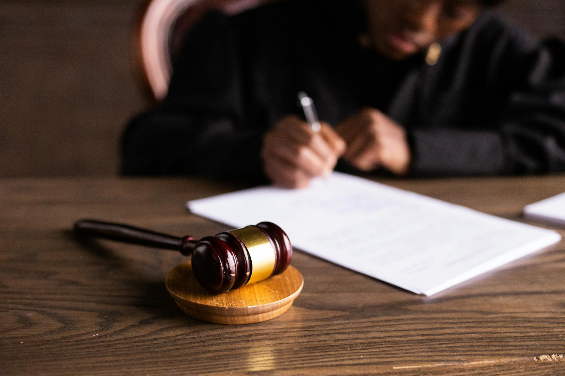 A gavel rests on a wooden table while a person in a dark robe writes on documents in the background.