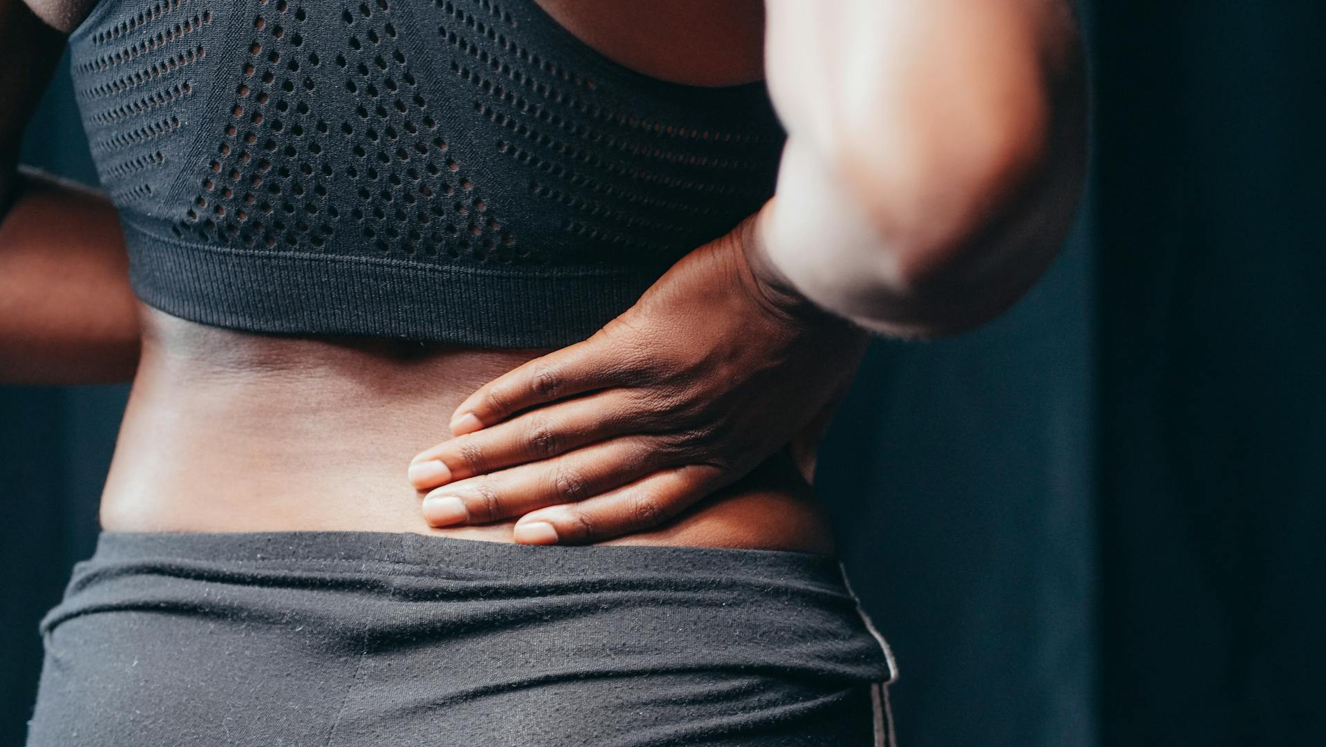 Person wearing black athletic clothing holds their lower back with one hand, suggesting discomfort or pain in the back area.