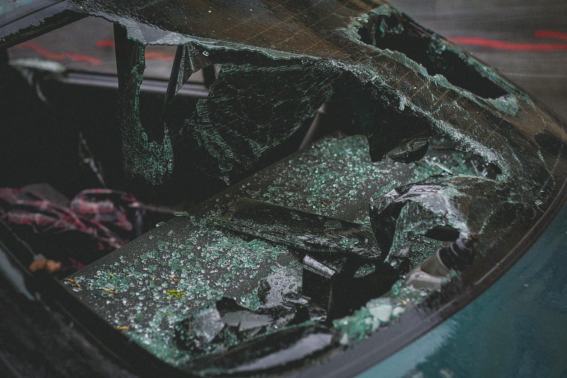 A shattered car windshield with broken glass covering the dashboard and front seats, indicating severe damage to the vehicle.