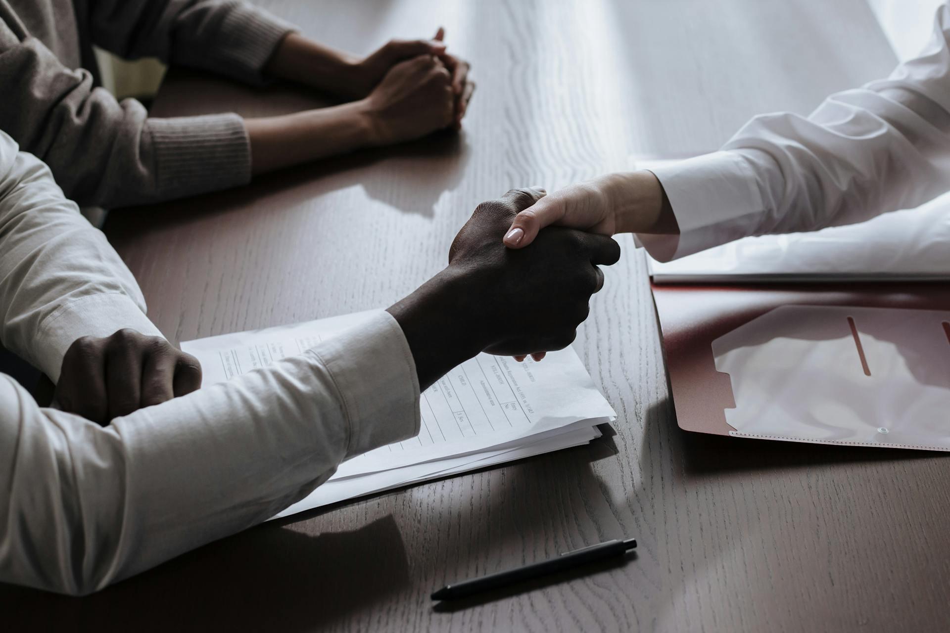 Two people shake hands over a desk with documents, while a third person sits nearby with hands clasped.