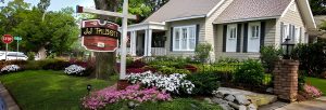 Well-maintained house with a front porch, surrounded by a landscaped yard with flowers and a sign reading "JJ THIBOT." A stop sign is visible at the street corner.