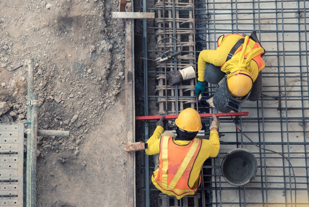 Two construction workers in yellow safety gear and hard hats are working with steel rebar and concrete on a building site, viewed from above.