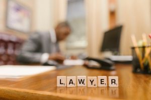 Scrabble tiles spelling "LAWYER" on a desk, with a person in a suit working at a desk in the blurred background.