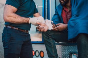 A person wearing gloves bandages another person's injured hand while sitting on the back of an emergency vehicle.