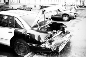 A damaged car with a severely crumpled and open trunk is parked on the street beside other vehicles.