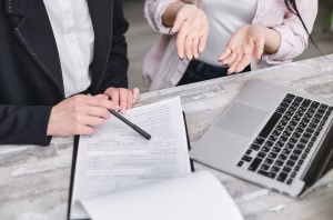 Two people sit at a desk with a laptop and paperwork; one points at a document with a pen, while the other gestures with open hands.