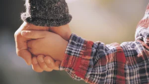 Close-up of two people holding hands, one wearing a gray sweater and the other wearing a red and blue plaid shirt.