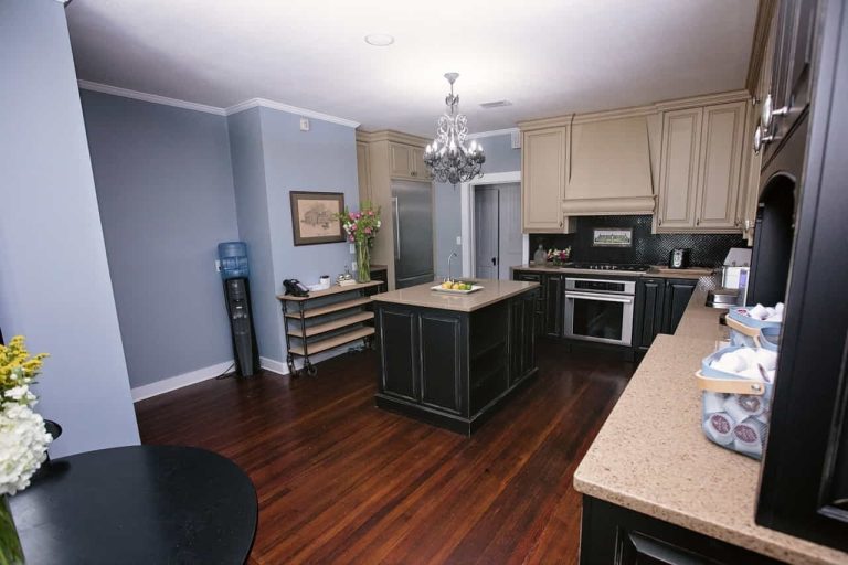 Modern kitchen with dark wood floors, beige cabinets, a central island, stainless steel appliances, and a chandelier; decorative items and a water cooler are also visible.