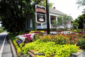 A law office sign for JJ Talbott stands in front of a house with a large, colorful flower garden along a roadside.