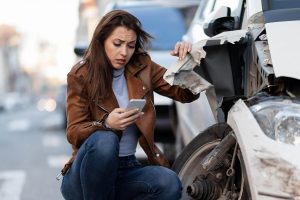 A woman crouches next to a damaged car, looking at her phone with a concerned expression after an apparent accident on a city street.
