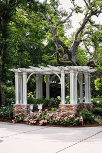 A white pergola with brick columns stands in a garden surrounded by green trees, flowering plants, and pink roses along a paved walkway.