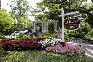 A law office sign reading "J.J. Talbott, Attorney at Law" stands by a garden with colorful flowers and greenery, with a pergola and parked cars in the background.