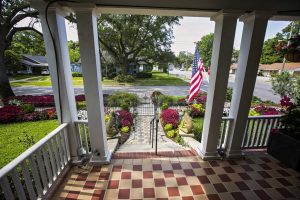 View from a tiled front porch overlooking a landscaped garden with flowers, a pathway, two stone lion statues, and an American flag, facing a suburban street.