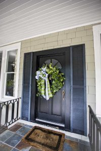 Black front door with a green wreath and plaid ribbon, flanked by dark shutters. A welcome mat lies in front on a tiled porch floor.