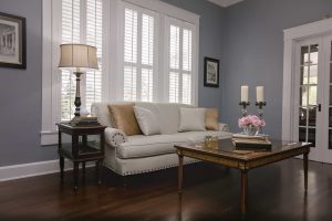 A living room with a white sofa, beige pillows, a side table with a lamp, a wooden coffee table with candles and flowers, and tall windows with white shutters.