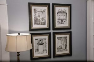 Four black-and-white architectural drawings in black frames are arranged in a grid on a gray wall next to a floor lamp with a white shade.