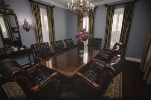 A formal dining room with a polished wooden table, eight leather chairs, a chandelier, tall windows with curtains, and a vase of flowers in the center.