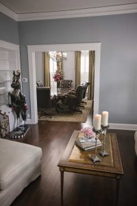 A view of a living room with a coffee table, candles, and books in the foreground, leading to a dining area with a table, chairs, and a chandelier in the background.