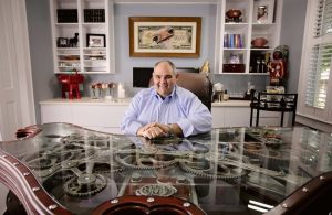 A man in a blue shirt sits at a glass-top desk with visible gears, in an office with shelves, framed art, and various decorations.