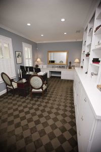 Home office with two vintage chairs at a desk, built-in white shelves, a large mirror, and checkered carpet. Room is well-lit with recessed ceiling lights and table lamps.