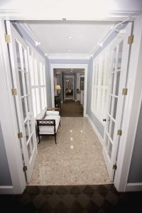 A hallway with glass double doors, marble tile flooring, a bench, and large windows with white blinds, leading to another room with carpet and a lit lamp.