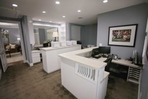 Reception area of an office with white desks, computer monitors, file organizers, and a sports-themed picture on the wall; lounge area is visible in the background.