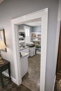 Small home office with shelves, a printer, various office supplies, and a window with white shutters, viewed from the doorway.
