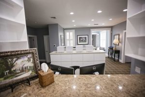 Reception area with a marble counter, tissue box, framed picture, two computer monitors, and white partitions. Behind are grey walls, artwork, and a side table with a lamp.