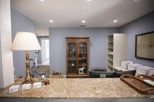 Reception desk with a marble countertop, lamp, baskets, and business cards in the foreground; cabinet, shelves, and desk in the background against light blue walls.