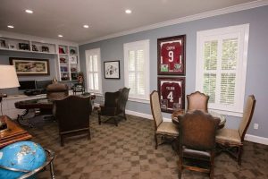Home office with a desk, computer, chairs, bookshelves, framed sports jerseys on the wall, windows with white shutters, and a globe on a side table.