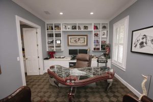 Home office with a glass-top wooden desk, leather chair, computer, built-in shelves with books and decor, and a framed dog drawing on a gray wall near a window with white shutters.