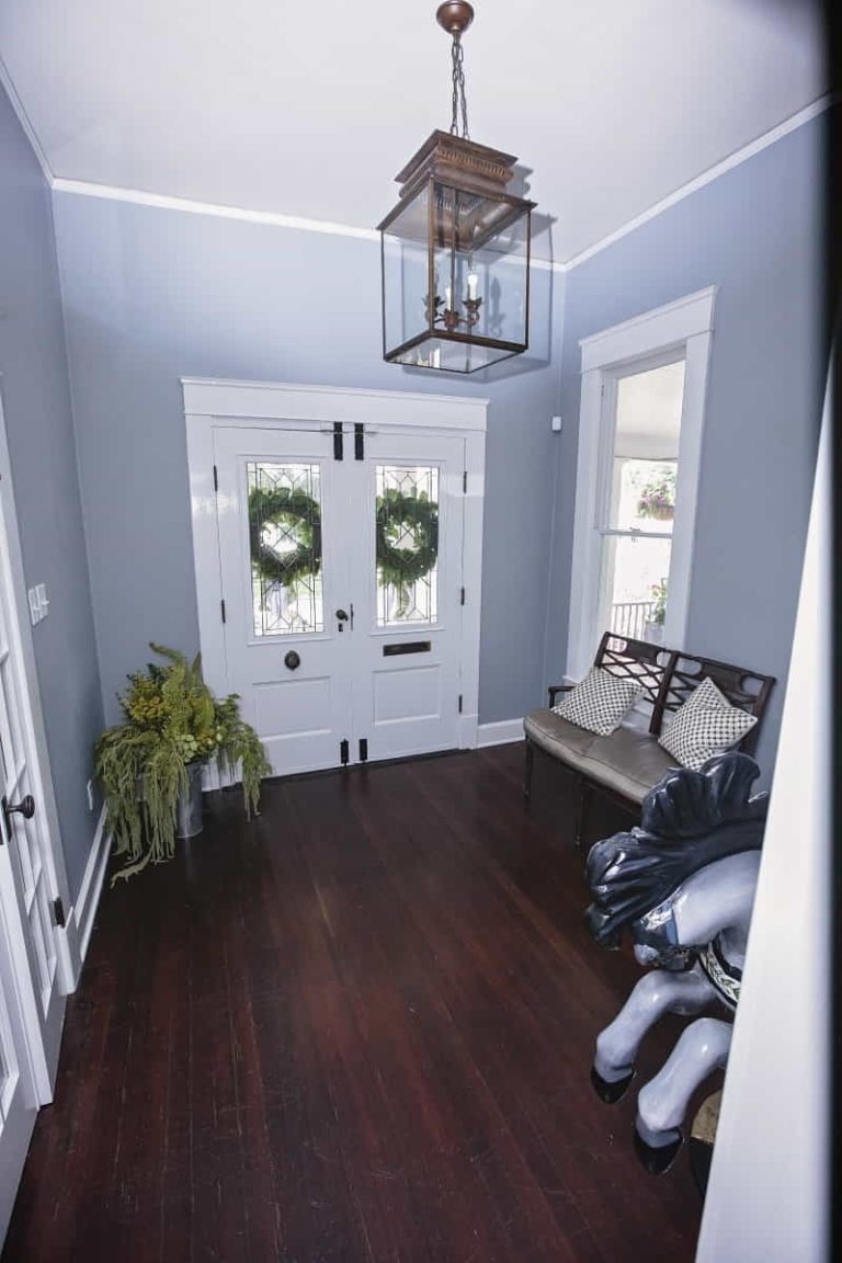 A hallway with dark wooden floors, blue-gray walls, a white double front door with wreaths, a bench with pillows, a plant, and a decorative horse sculpture.