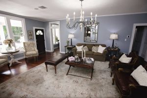 A spacious living room with blue walls, a beige sofa, armchairs, a large rug, a coffee table with candles, and a chandelier, with natural light from windows.