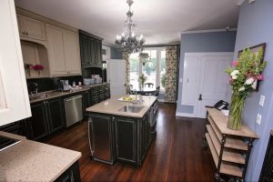 Modern kitchen with dark cabinets, central island with sink, stainless steel appliances, and a chandelier. There are flowers on the countertop and side table, and a window in the background.