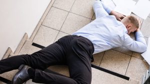 A man in business attire lies face down on stairs with scattered papers around him, appearing to have fallen.