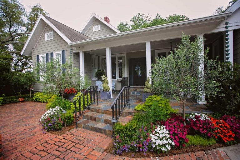 A single-story house with a front porch, white columns, brick walkway, and colorful flower beds along the entrance steps.