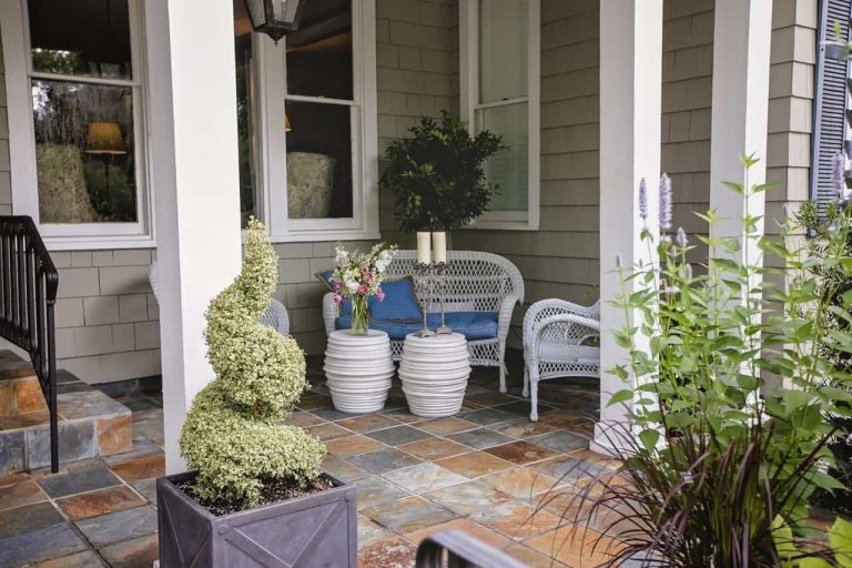 Covered porch with wicker chairs, round white tables with flowers, potted plants, and slate tile flooring near large windows.