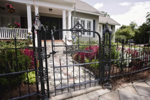 A black wrought iron gate opens to a walkway leading to the front porch of a gray house with white columns, surrounded by bushes and flowers.