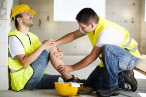 Two construction workers wearing safety vests and helmets; one sits on the floor holding his injured knee while the other checks the injury.