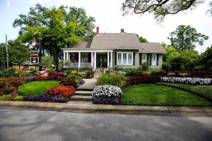 A single-story house with a porch, surrounded by colorful flowerbeds, manicured lawns, and trees, viewed from across the street.